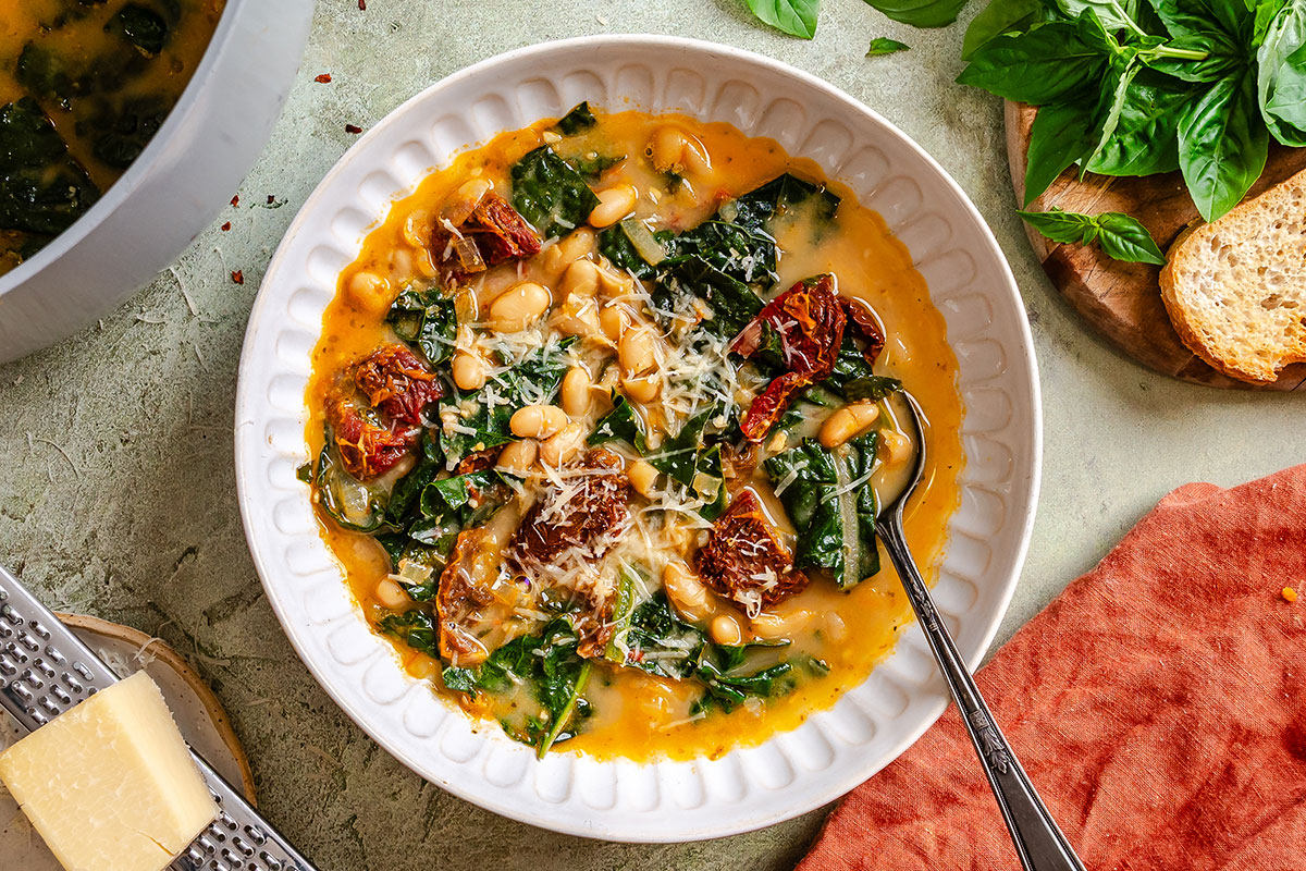 A bowl of white bean and kale soup topped with grated cheese and sun-dried tomatoes, with a spoon in the bowl and bread, basil, and cheese nearby.