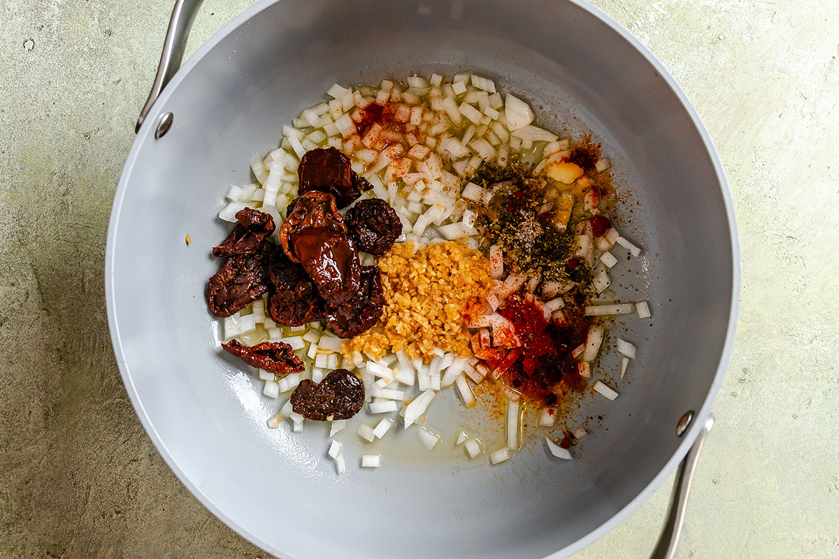 Chopped onions, garlic, sun-dried tomatoes, oil, and spices in a light gray pan on a light surface, ready to be cooked.