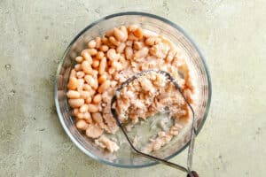 A glass bowl filled with cannellini beans, some whole and some partially mashed with a metal potato masher, sits on a light green textured surface.