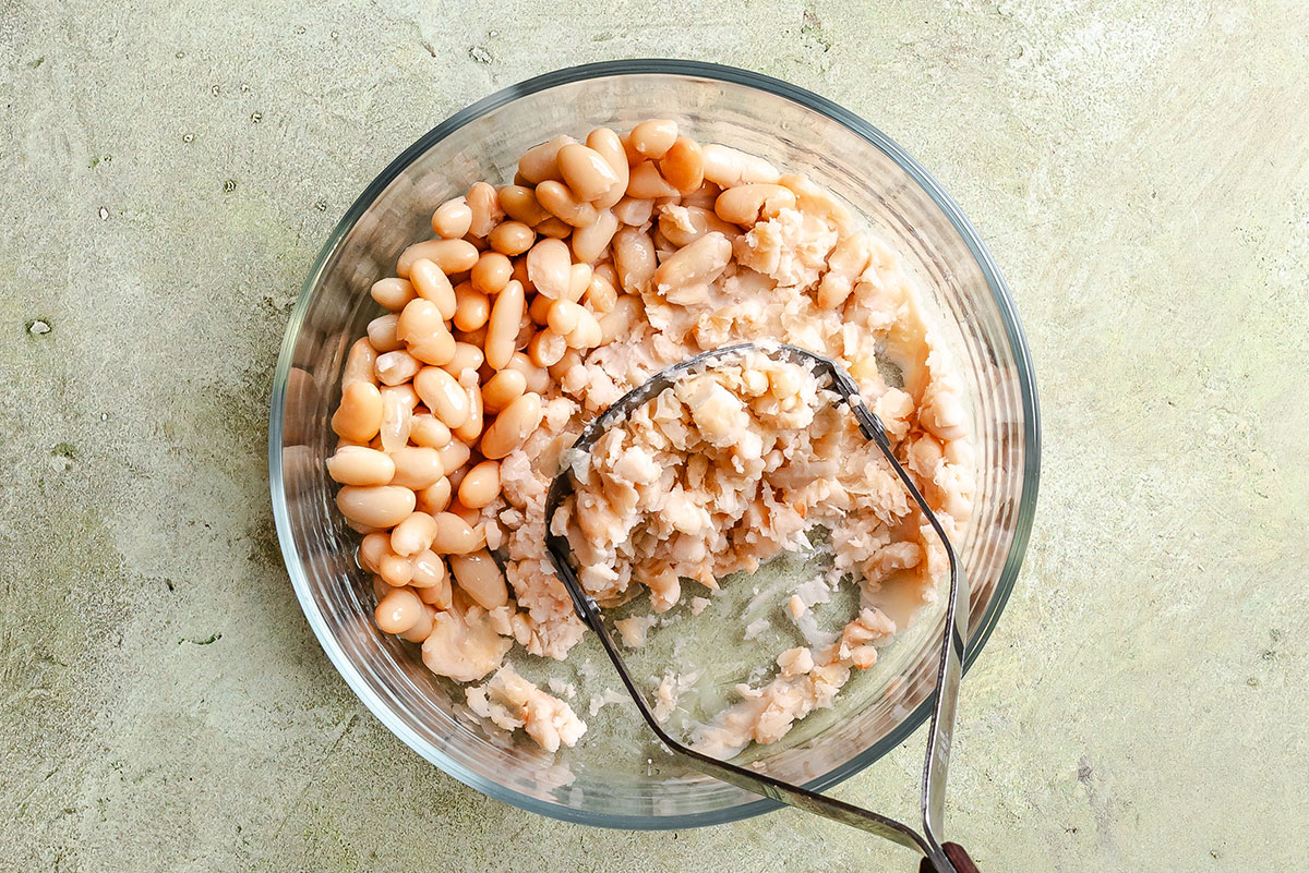 A glass bowl filled with cannellini beans, some whole and some partially mashed with a metal potato masher, sits on a light green textured surface.