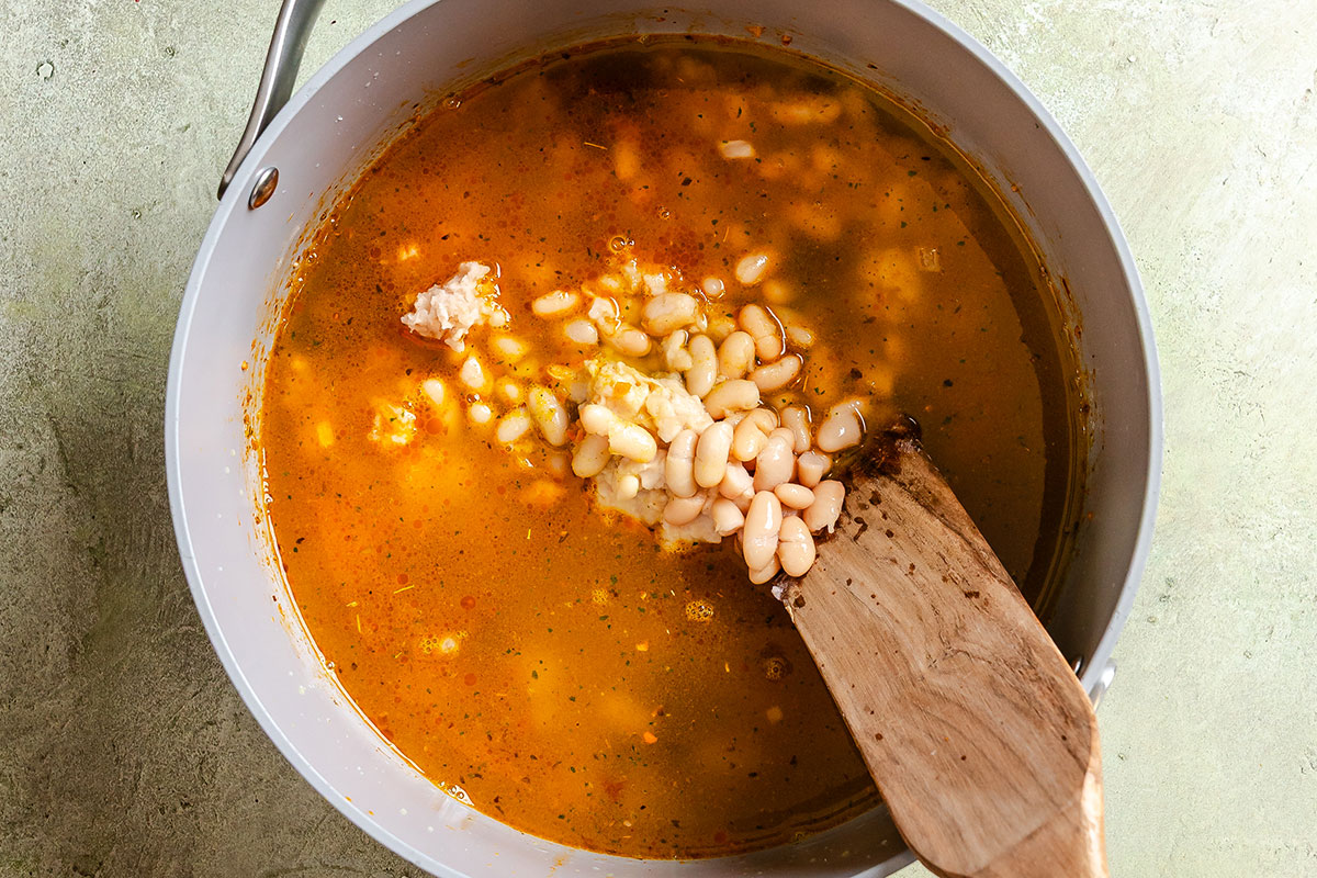 A pot of orange broth with white beans being stirred by a wooden spatula.