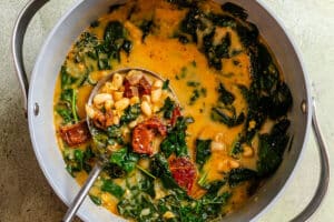 A pot of soup containing white beans, chopped kale, sun-dried tomatoes, and broth with a ladle resting inside.