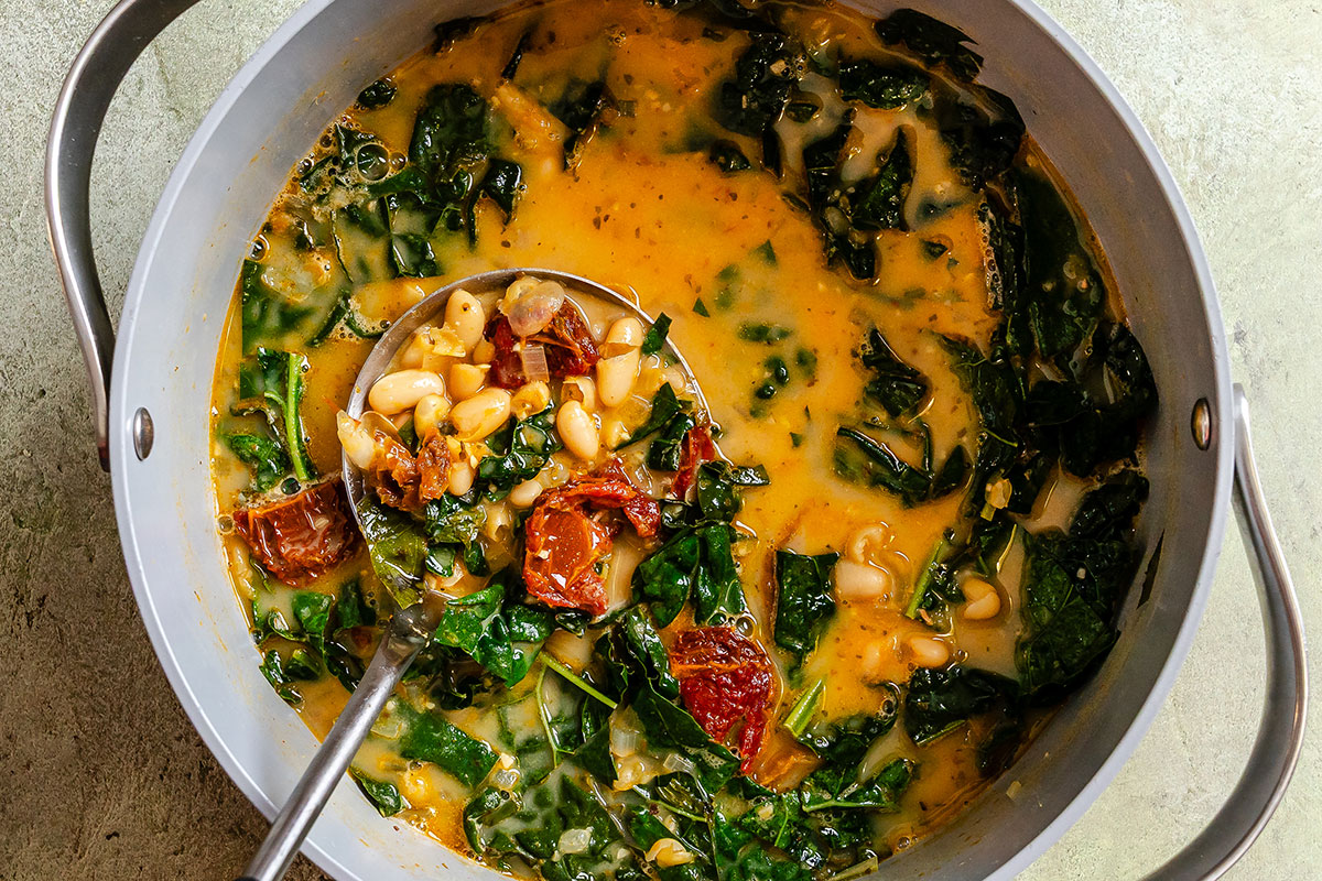 A pot of soup containing white beans, chopped kale, sun-dried tomatoes, and broth with a ladle resting inside.