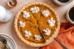 Pumpkin pie sliced into eight pieces, each topped with whipped cream, sits on a table with a pie server, mugs, plates, forks, and an orange napkin.