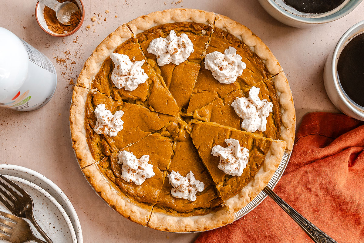Pumpkin pie sliced into eight pieces, each topped with whipped cream, sits on a table with a pie server, mugs, plates, forks, and an orange napkin.