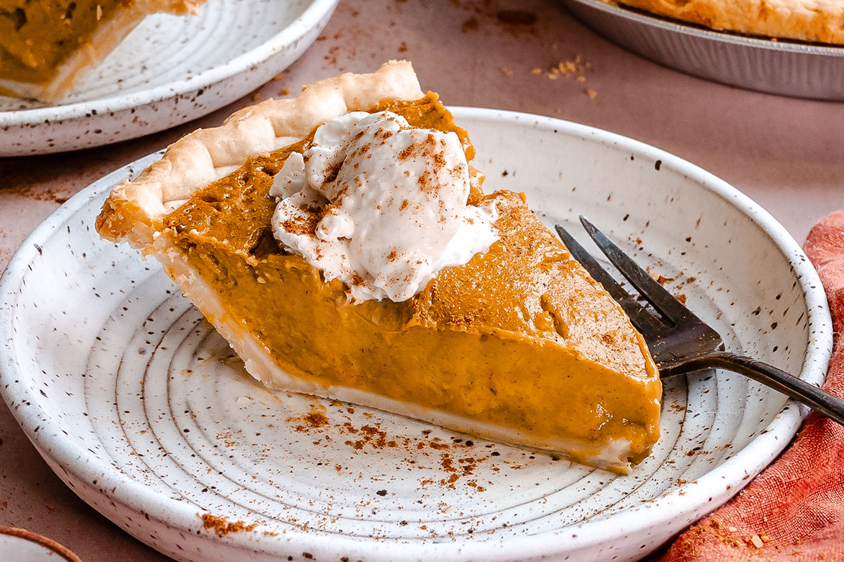 A slice of pumpkin pie topped with whipped cream and a sprinkle of cinnamon sits on a speckled plate with a fork beside it.