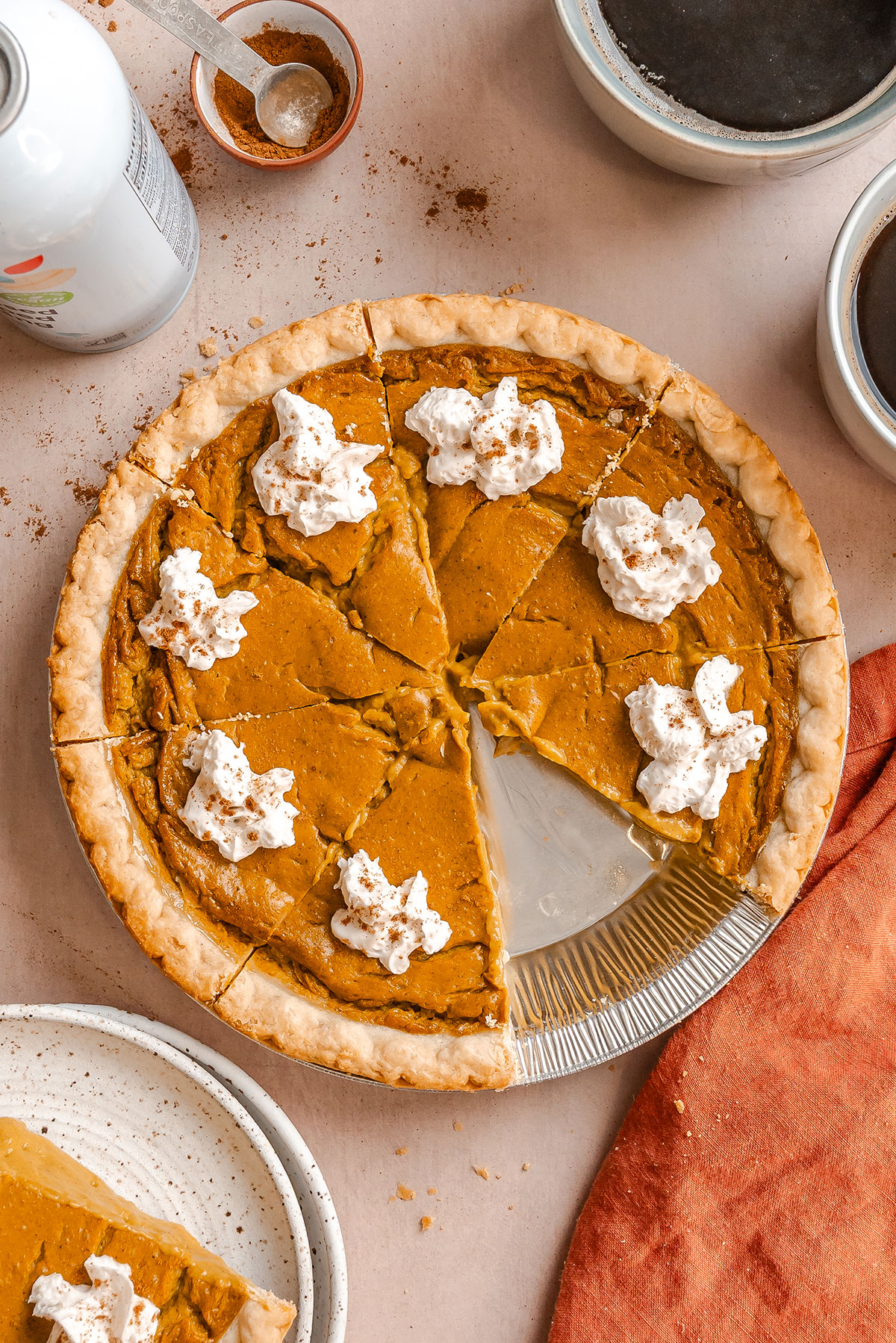 Pumpkin pie with a missing slice, topped with whipped cream dollops, sits on a table with coffee cups, a can, a plate with pie, and a measuring spoon.