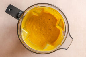 Top view of a blender filled with thick, smooth yellow-orange liquid, likely a blended soup or sauce, on a light-colored surface.