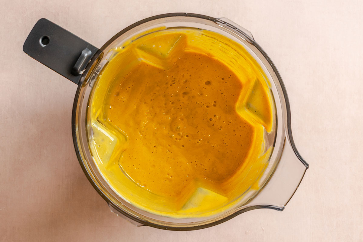 Top view of a blender filled with thick, smooth yellow-orange liquid, likely a blended soup or sauce, on a light-colored surface.