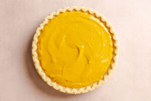 Unbaked pie crust filled with smooth yellow pumpkin pie filling, viewed from above on a light surface.