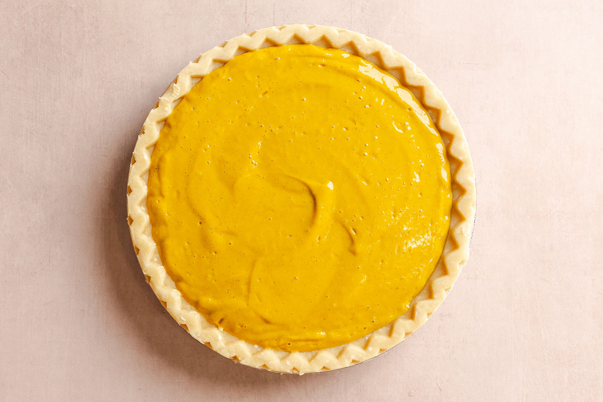 Unbaked pie crust filled with smooth yellow pumpkin pie filling, viewed from above on a light surface.