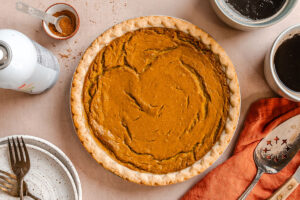 Pumpkin pie in a pie tin on a table, surrounded by a plate with forks, a pie server, a napkin, a small bowl of cinnamon, and two mugs of dark liquid.