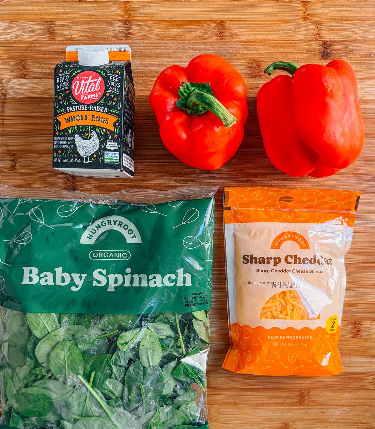 Carton of eggs, two red bell peppers, bag of organic baby spinach, and a bag of shredded sharp cheddar cheese on a wooden surface.