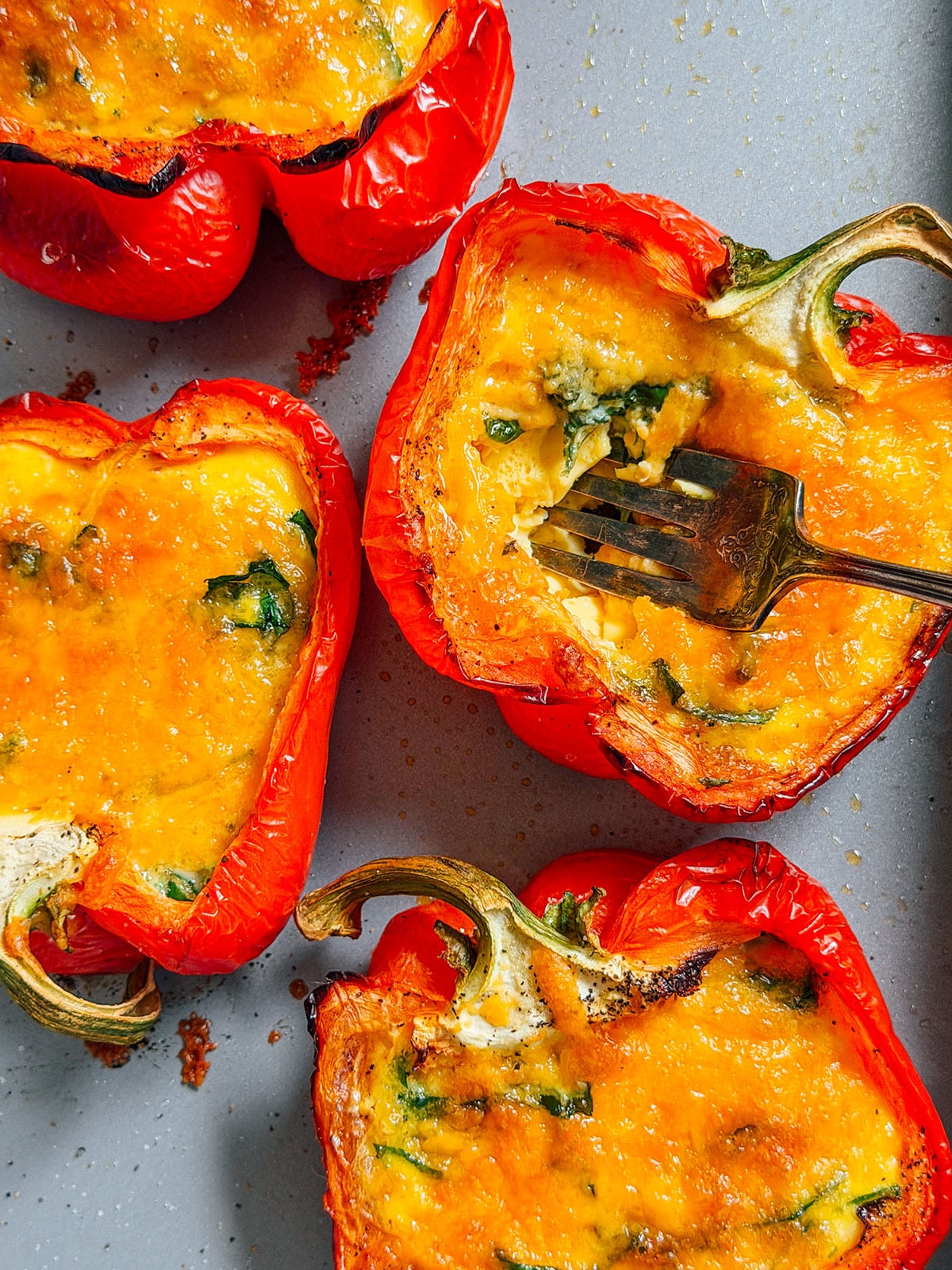 Four red bell pepper halves filled with a baked cheesy mixture sit on a gray surface. A fork rests in one pepper, revealing a cheesy, slightly browned filling with greens.
