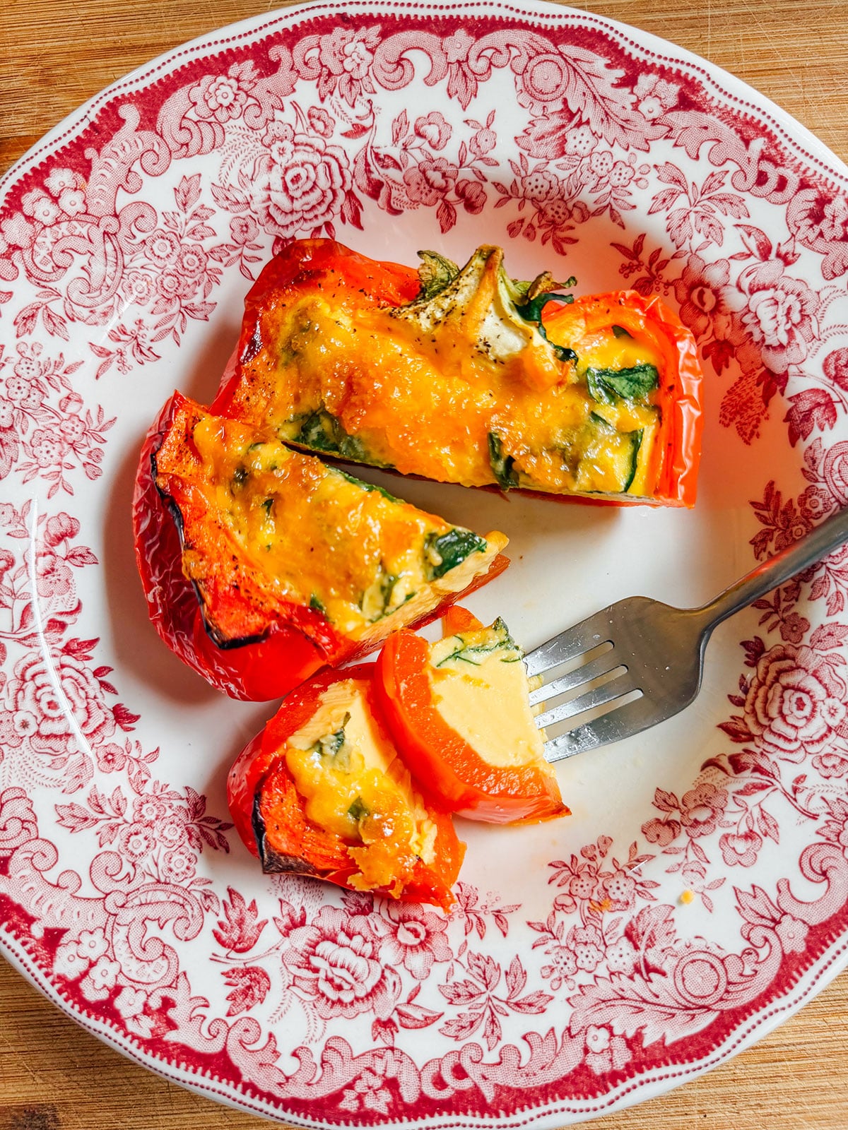 A stuffed red bell pepper filled with an egg and spinach mixture sits on a red and white floral plate, partly cut with a fork.