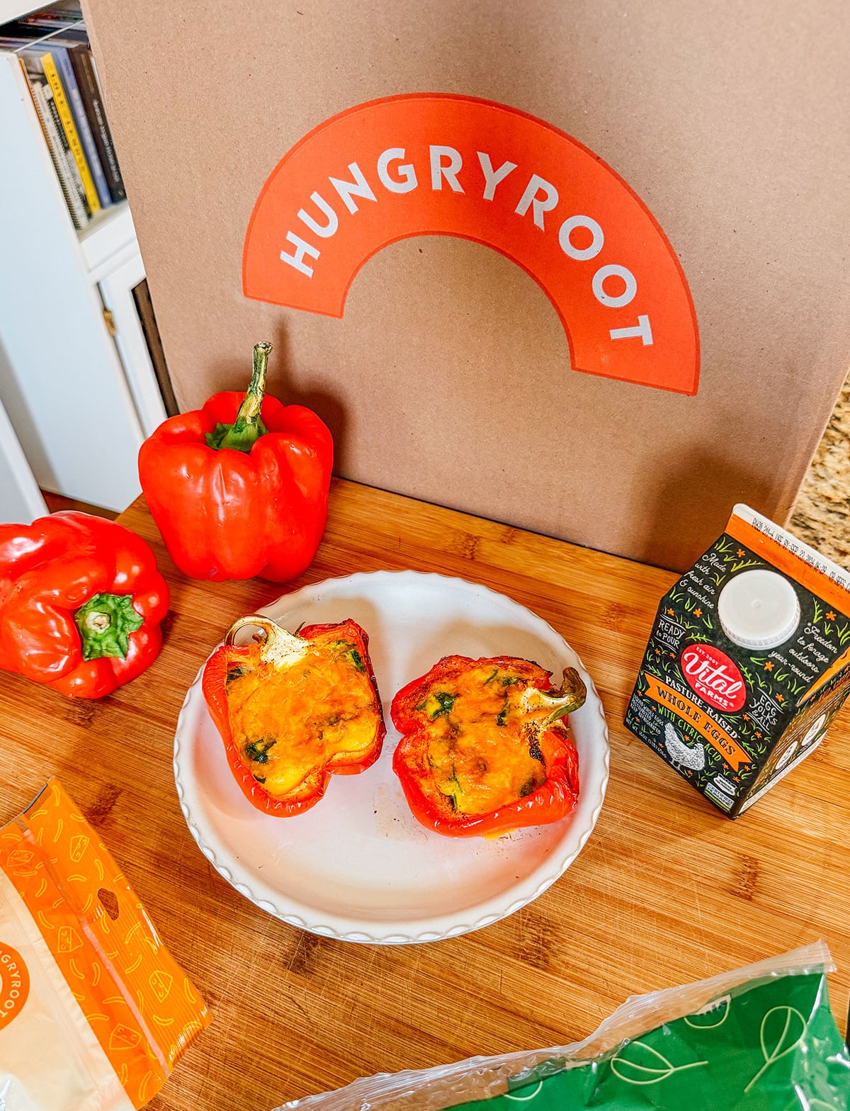A sliced stuffed red bell pepper on a plate sits on a wooden counter with whole peppers, oat milk, packaging, and a Hungryroot box in the background.