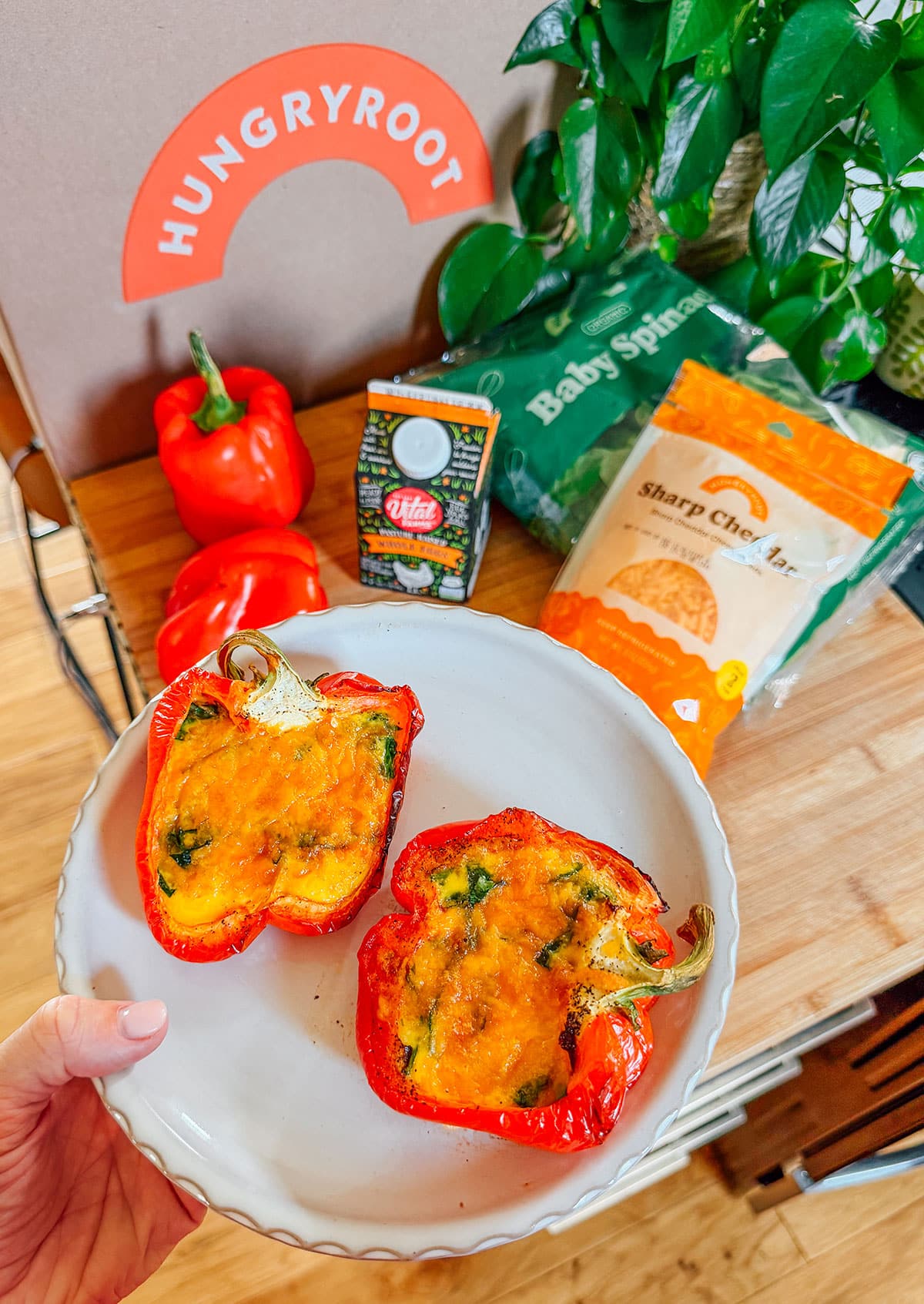 A hand holds a plate with two cooked stuffed red bell peppers. In the background are a Hungryroot box, red peppers, baby spinach, cheddar cheese, and a carton of milk.