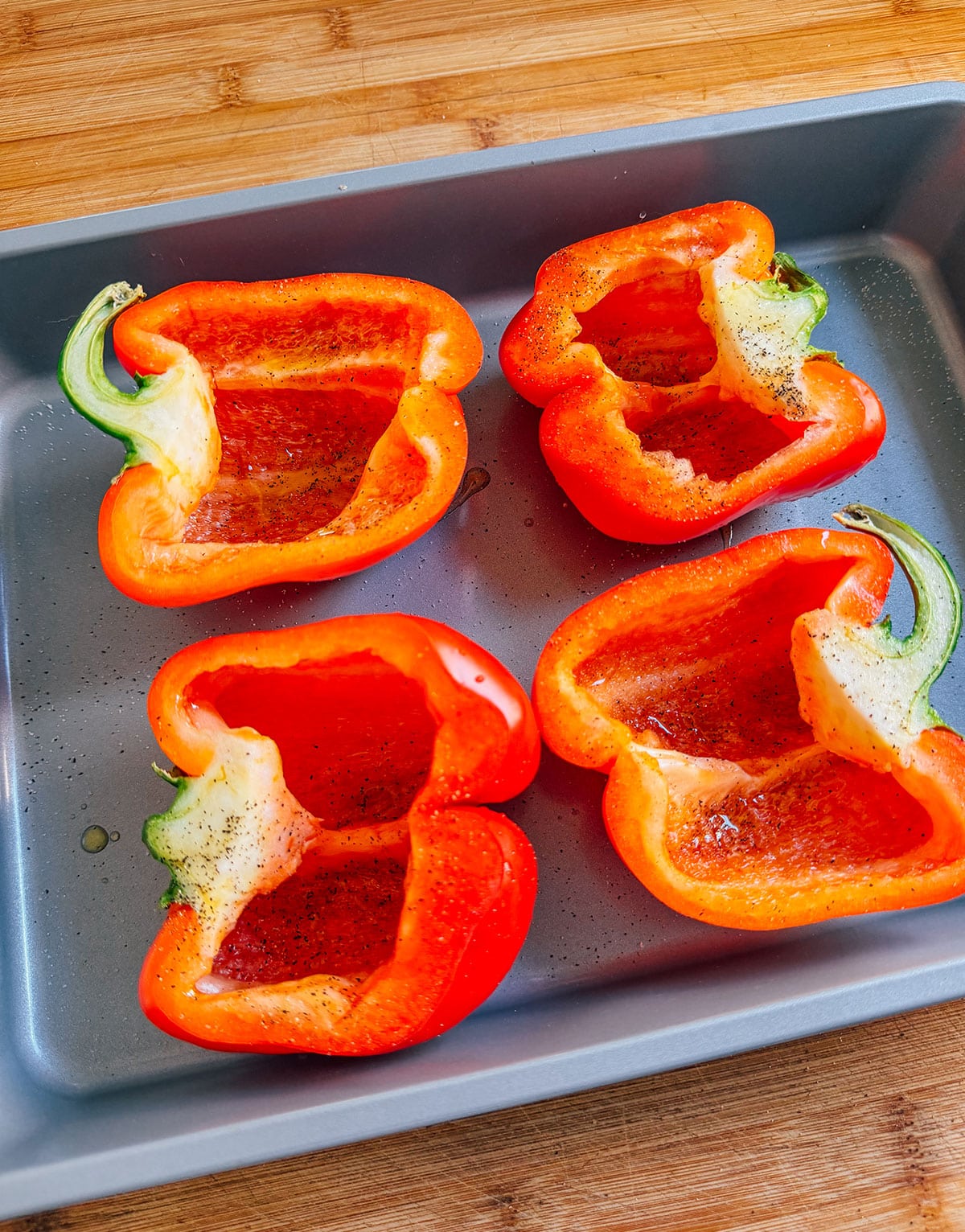 Four red bell pepper halves placed cut side up in a metal baking pan, seasoned with black pepper and oil, on a wooden surface.