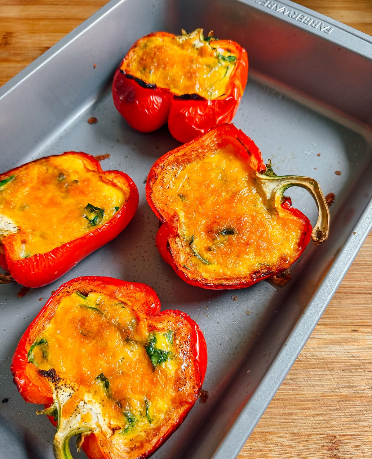 Four stuffed red bell pepper halves with melted cheese and herbs on top are arranged in a metal baking tray on a wooden surface.