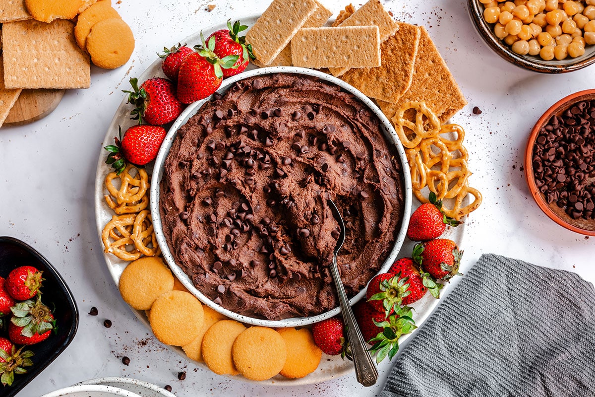 A bowl of brownie batter chocolate hummus topped with chocolate chips, surrounded by strawberries, graham crackers, pretzels, and vanilla wafer cookies.