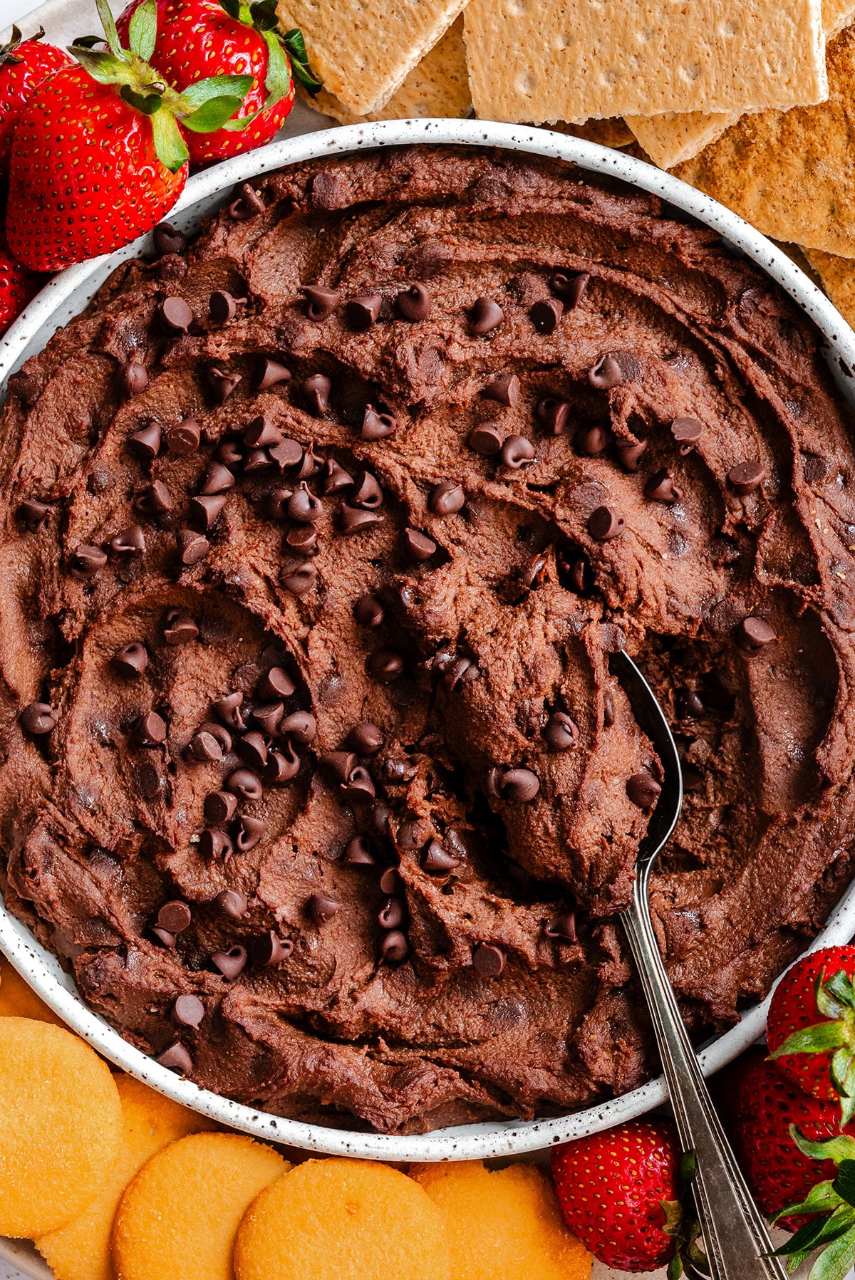 A bowl of chocolate chip dessert hummus with a serving spoon, surrounded by fresh strawberries and crackers.