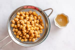 A metal strainer filled with chickpeas next to a small bowl of aquafaba, ready to be used for chocolate hummus.