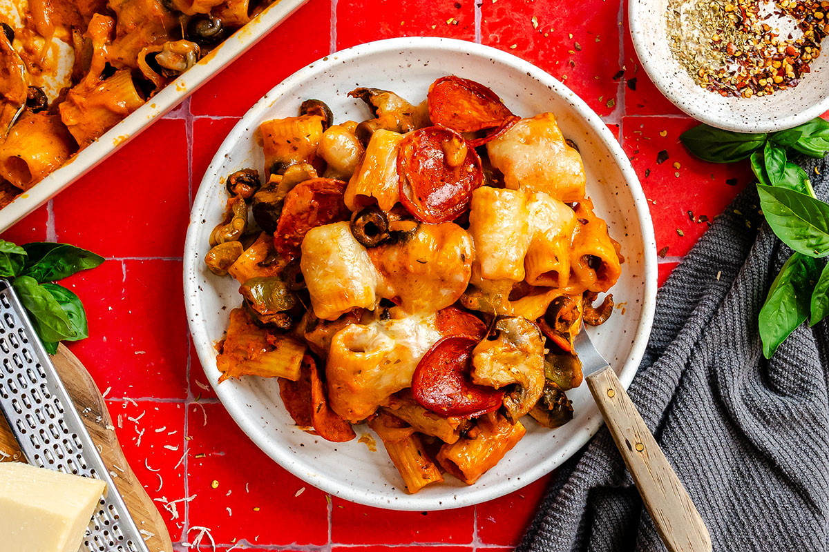 A plate of baked rigatoni pasta with melted cheese, pepperoni slices, and olives, with a small bowl of seasoning and a grater nearby on a red tiled surface.