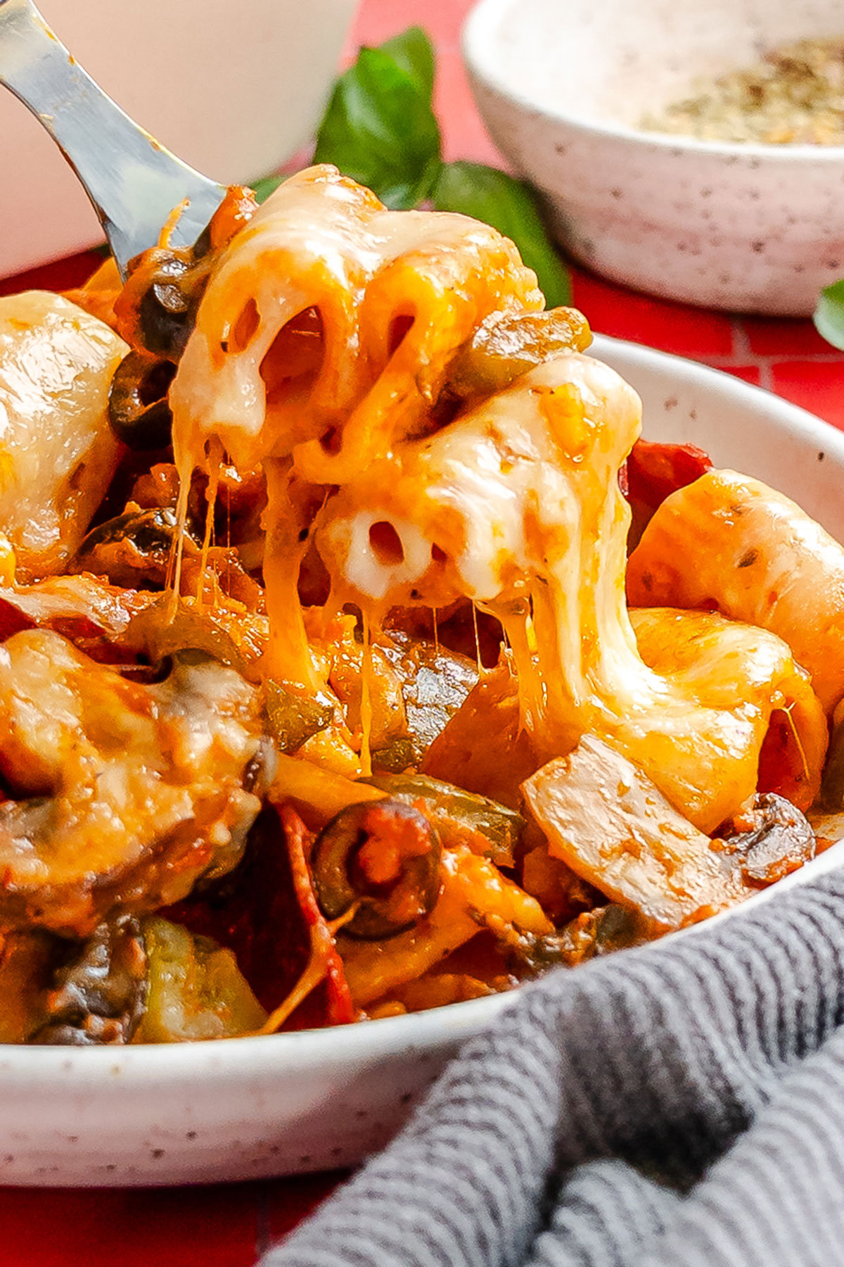 A fork lifting rigatoni pasta with melted cheese, olives, and vegetables from a bowl.