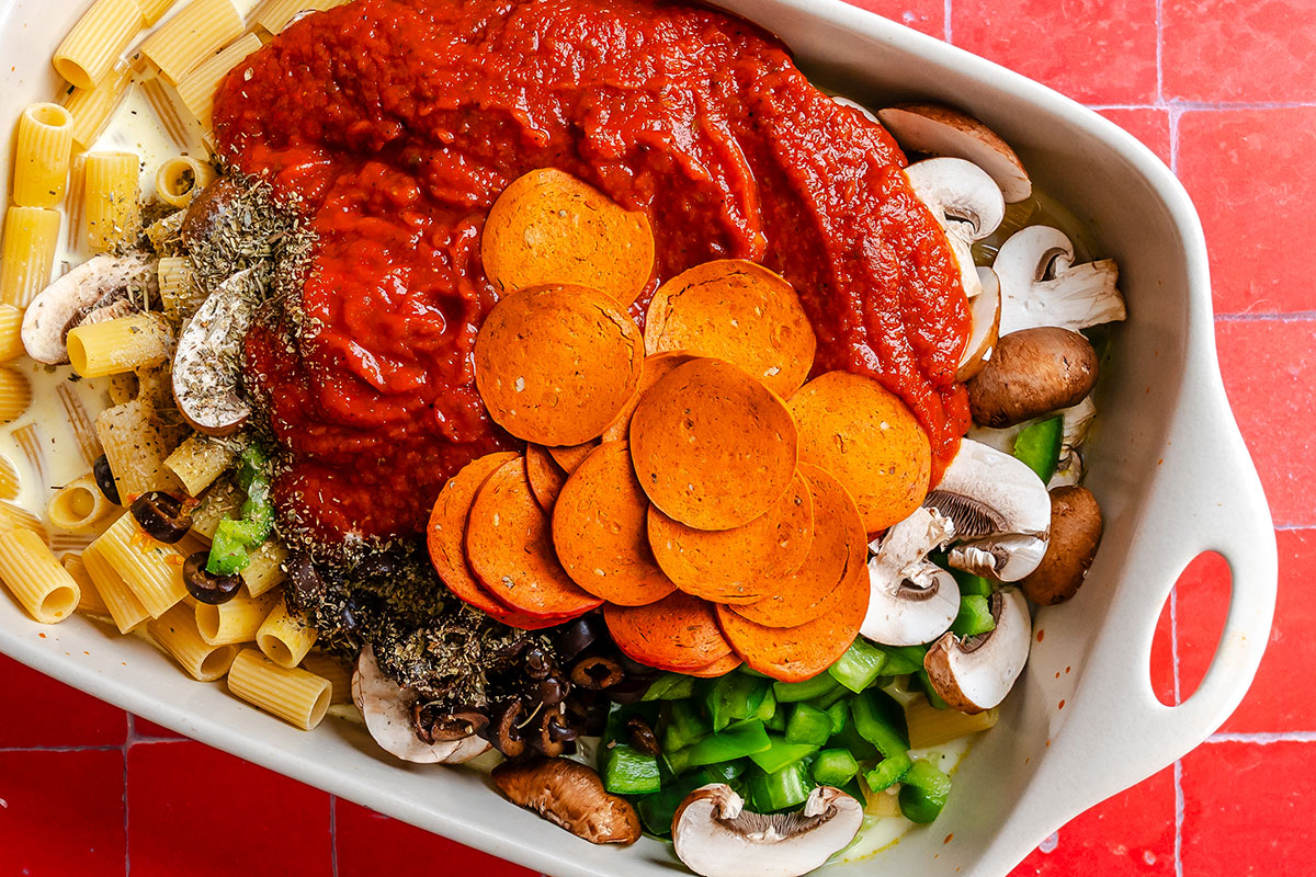 A baking dish filled with rigatoni, red sauce, sliced pepperoni, chopped green peppers, sliced mushrooms, and black olives.