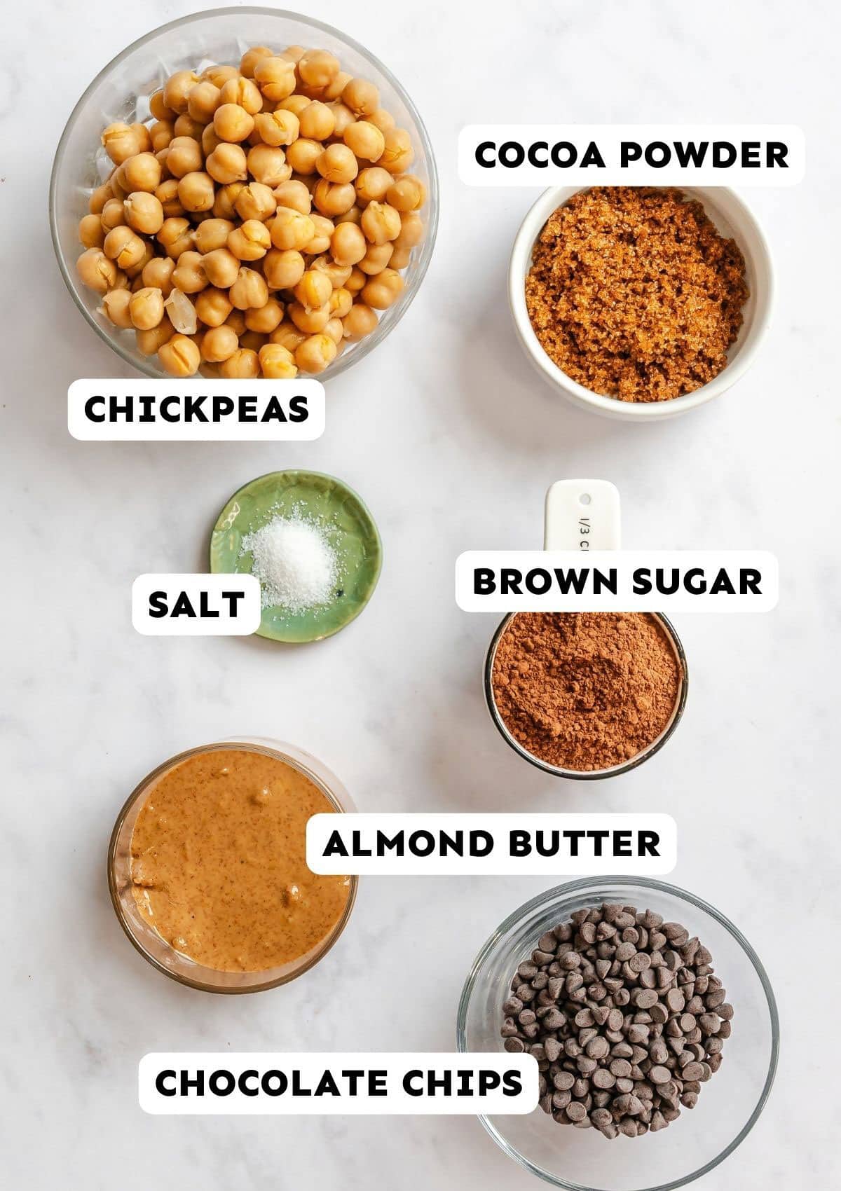 Ingredients for a chocolate hummus brownie batter recipe: chickpeas, cocoa powder, salt, brown sugar, almond butter, and chocolate chips, each with a corresponding label.