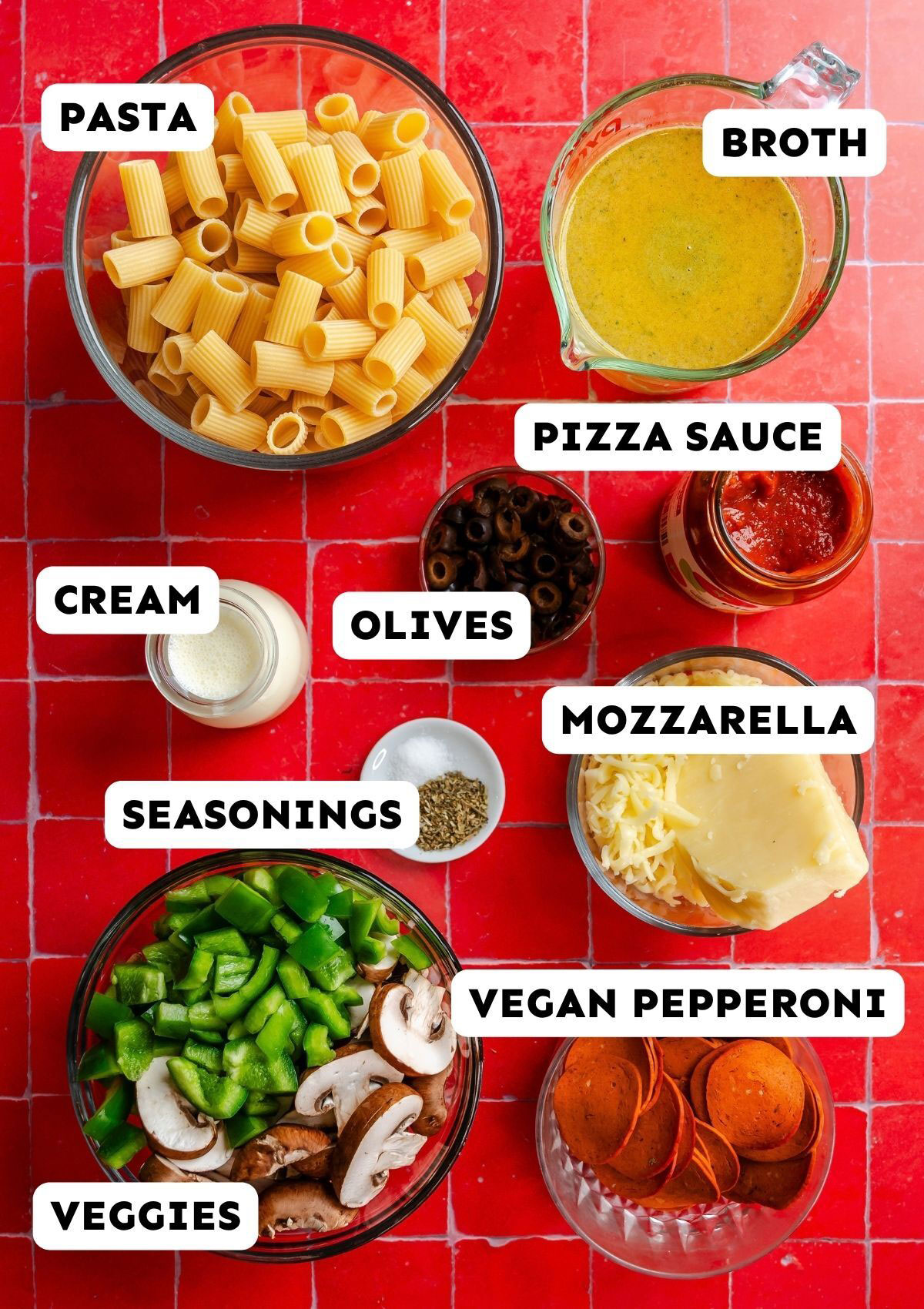 Ingredients for a recipe laid out on a red tiled surface, labeled as pasta, broth, pizza sauce, cream, olives, seasonings, mozzarella, vegan pepperoni, and veggies.