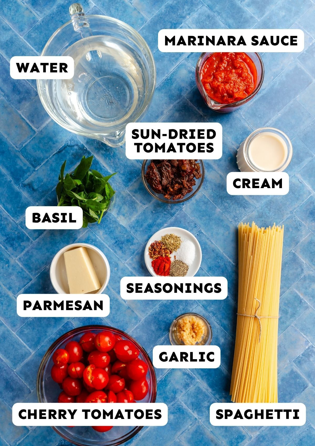 Ingredients for creamy tuscan Marry Me Pasta on a blue tile background, including water, marinara sauce, sun-dried tomatoes, cream, basil, seasonings, parmesan, garlic, cherry tomatoes, and spaghetti.