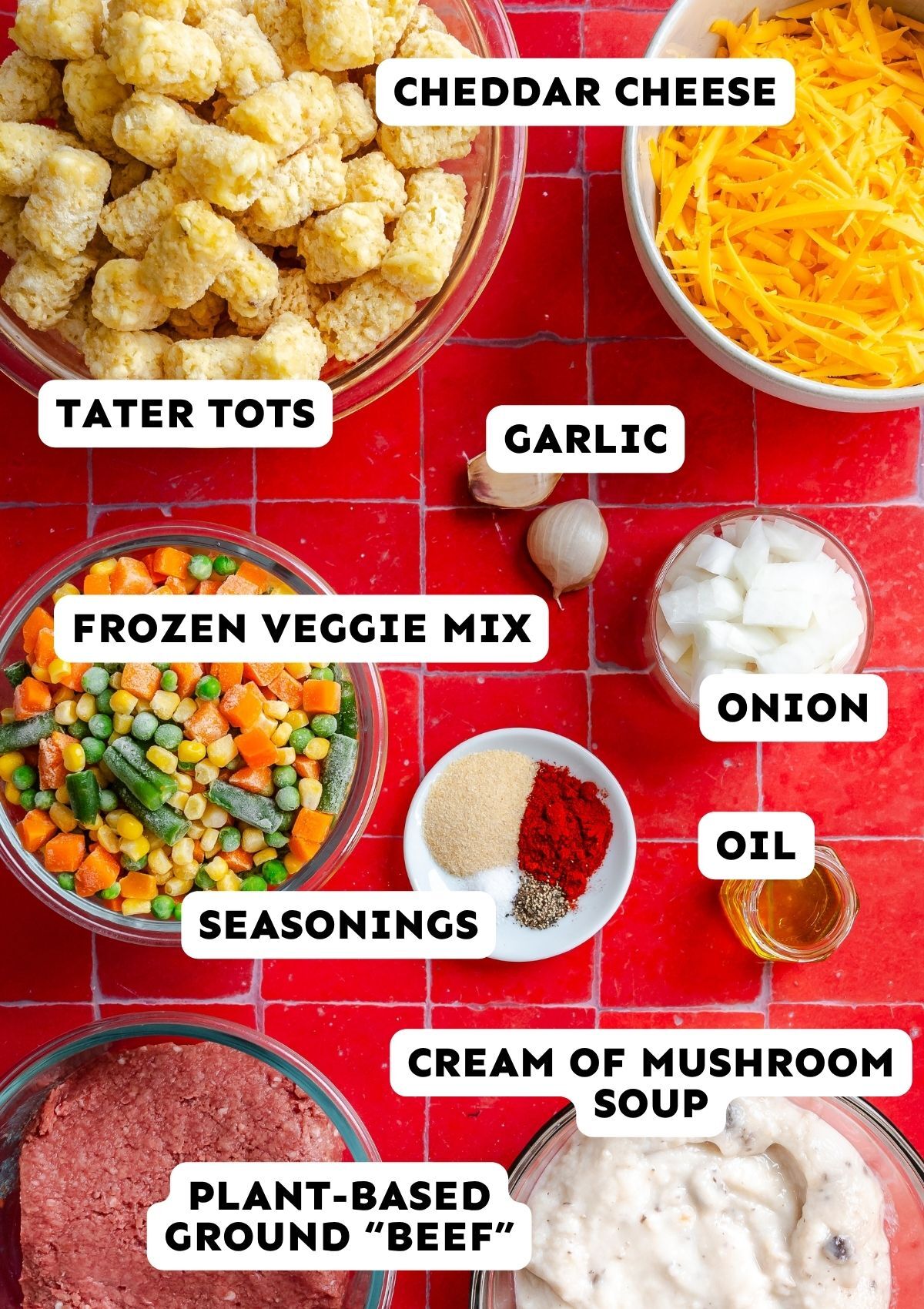 Overhead view of ingredients for a vegetarian tater tot casserole, including tater tots, cheddar, aromatics, frozen vegetables, spices, oil, plant-based beef, and cream of mushroom soup.