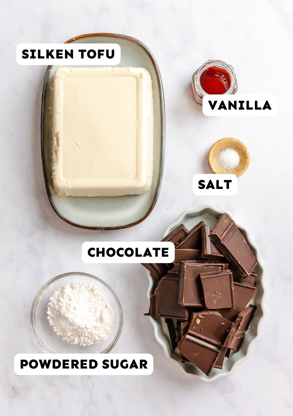 Ingredients for a recipe labeled silken tofu, vanilla, salt, chocolate, and powdered sugar arranged on a marble surface.