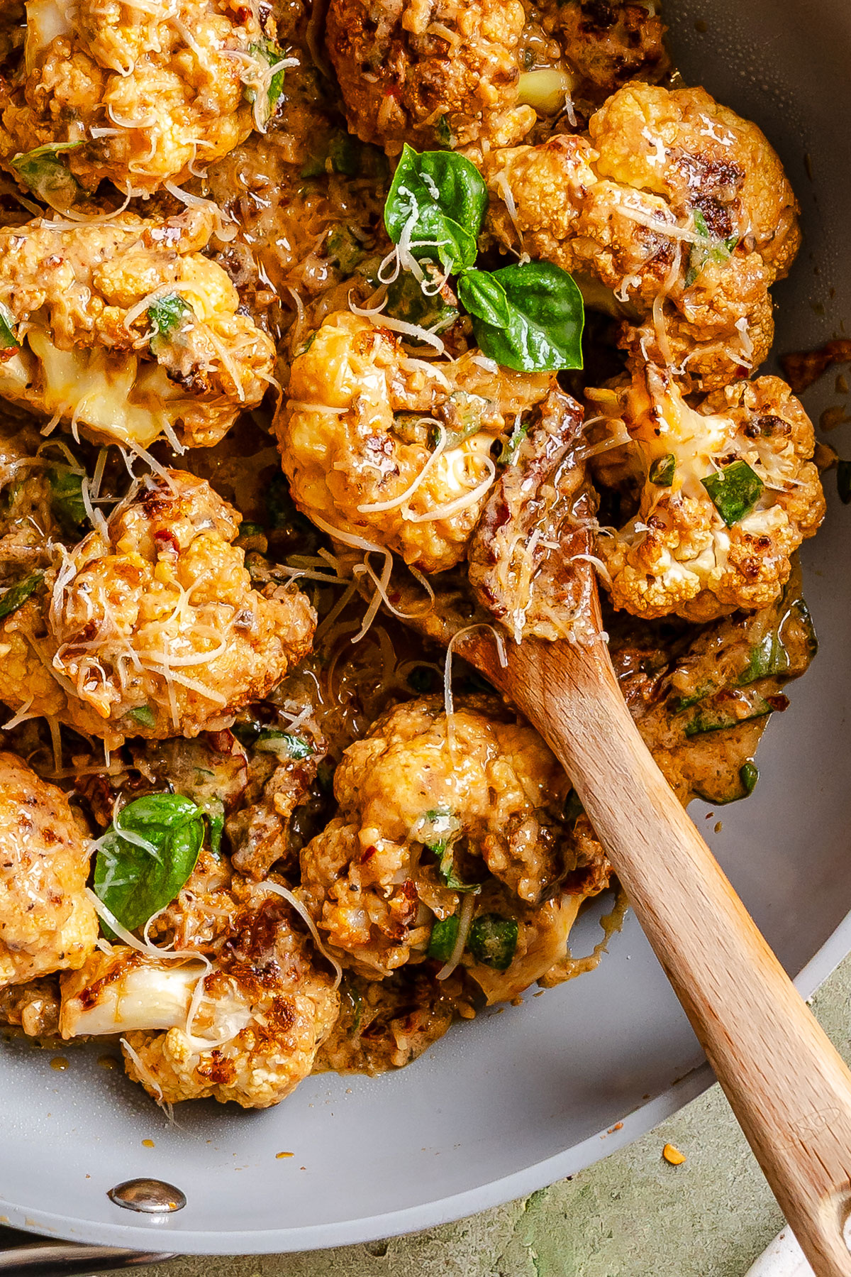 Roasted cauliflower florets in a creamy sauce with basil and grated cheese, being served with a wooden spoon in a pan.