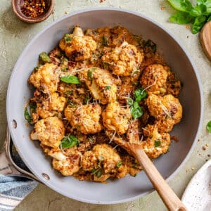 Pan-seared cauliflower florets in a creamy sauce, garnished with basil and shredded cheese, with a wooden spoon in the pan.