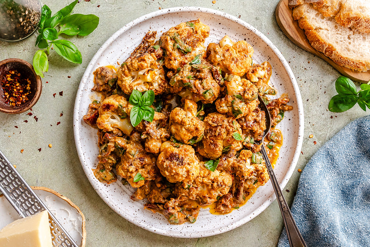 Roasted cauliflower florets in a creamy sauce garnished with fresh basil on a ceramic plate, served with bread and parmesan cheese.