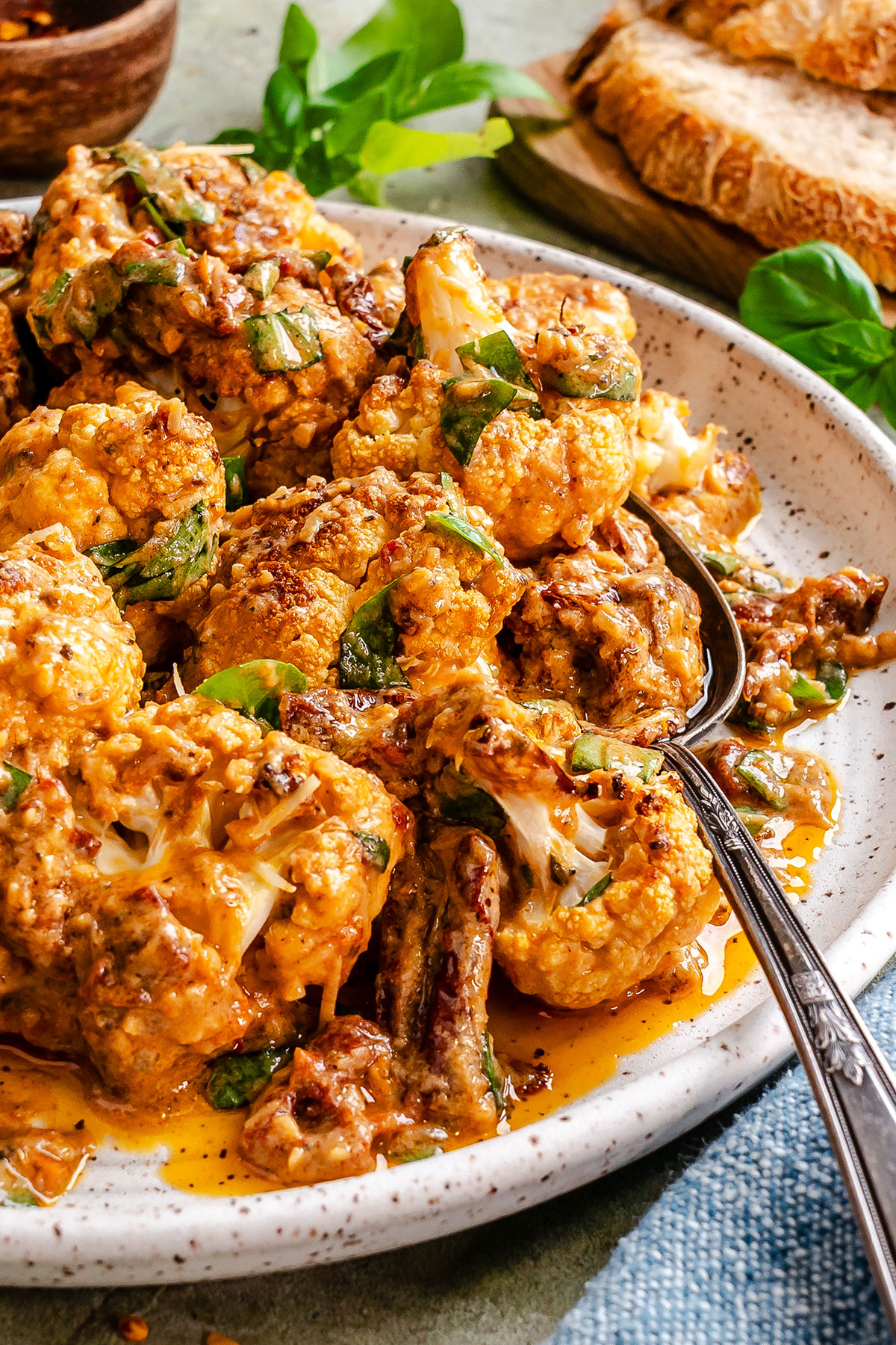 Roasted cauliflower florets coated in a creamy, red-tinted sauce with herbs, served on a speckled plate with a metal spoon.