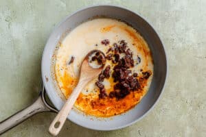 A wooden spoon stirs sun-dried tomatoes and cream in a grey skillet.