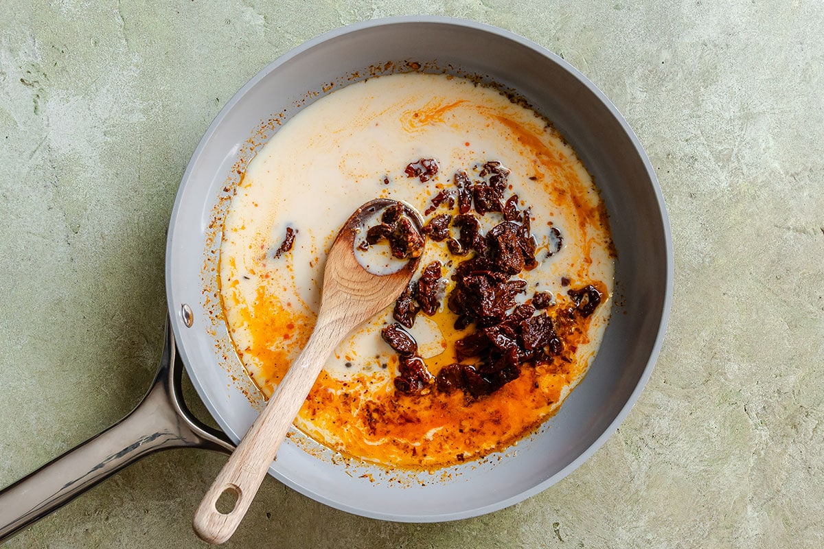 A wooden spoon stirs sun-dried tomatoes and cream in a grey skillet.