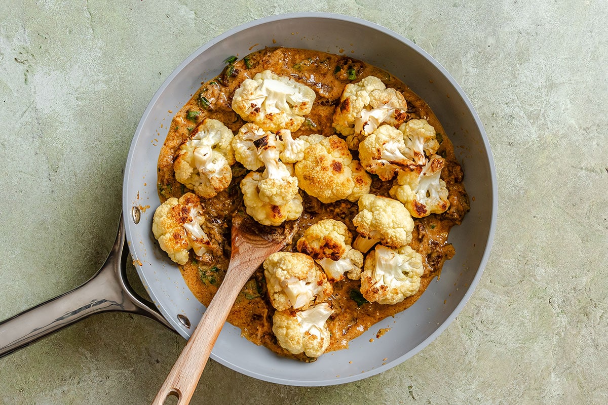 Roasted cauliflower florets in a creamy orange sauce in a skillet with a wooden spoon.