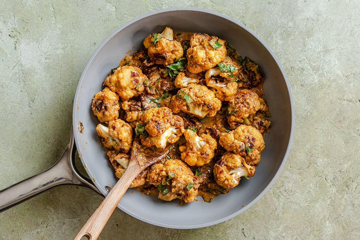 Roasted cauliflower florets in a creamy sauce with herbs, served in a skillet with a wooden spoon.