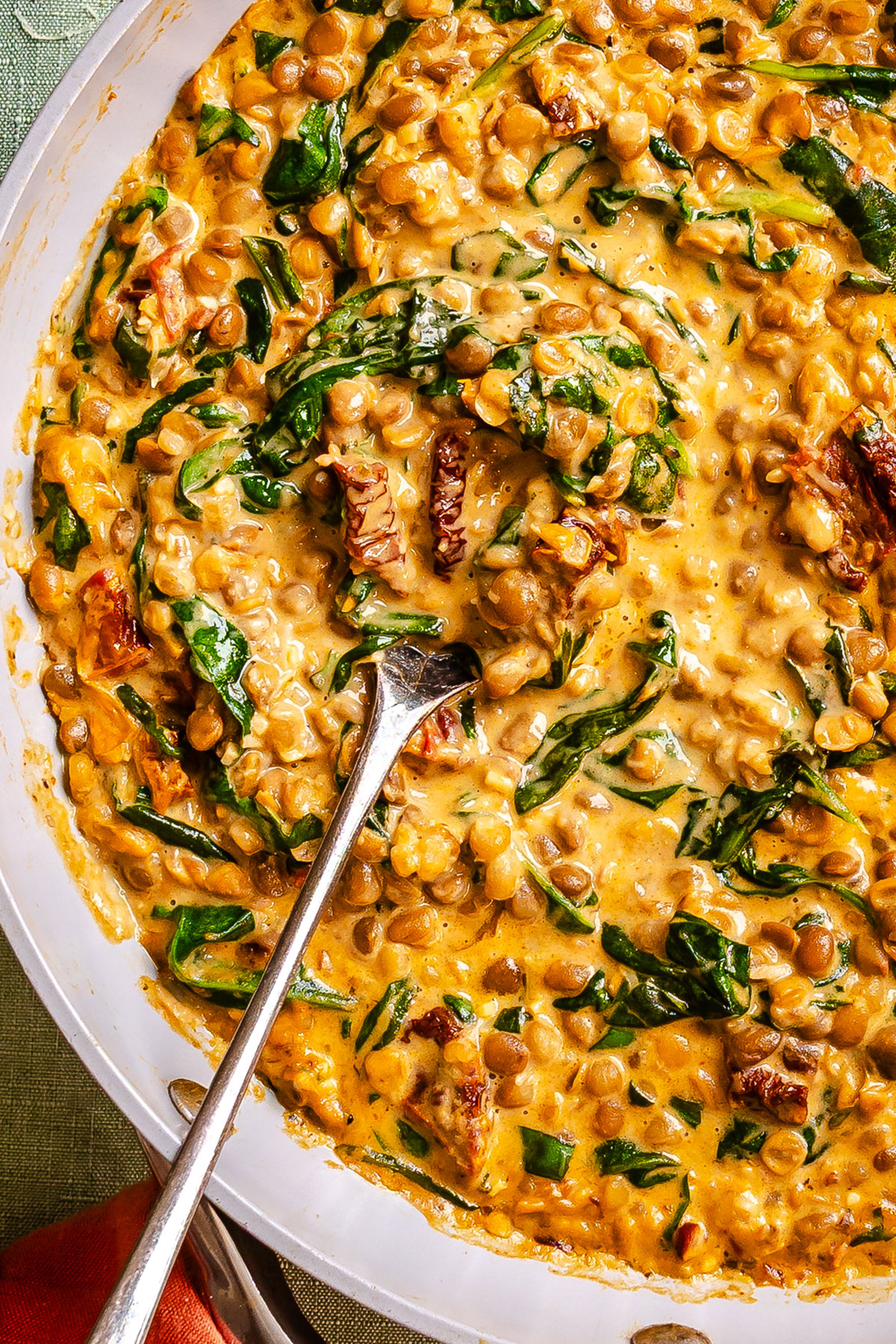 Creamy lentil stew with spinach and sun-dried tomatoes in a skillet with a metal spoon.