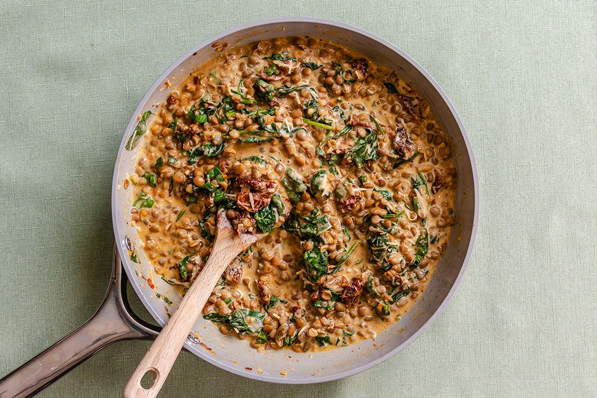Creamy lentils with spinach and sun-dried tomatoes in a skillet with a wooden spoon.