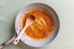 A wooden spoon stirring toasted spices and oil in a light-colored skillet.