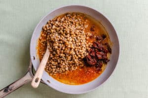 A light-colored pan containing cooked lentils and sun-dried tomatoes in oil, with a wooden spoon resting in the center.