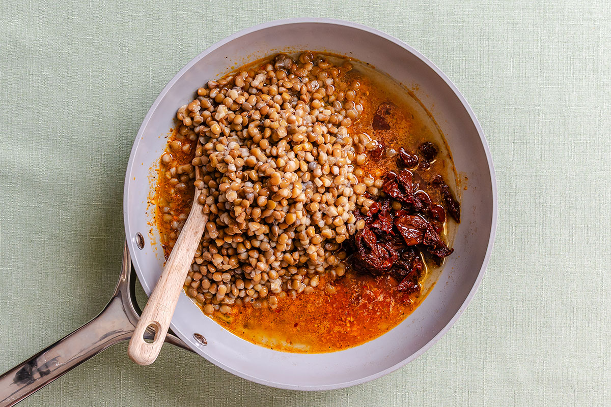 A light-colored pan containing cooked lentils and sun-dried tomatoes in oil, with a wooden spoon resting in the center.