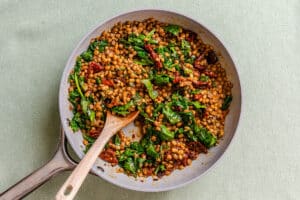 A skillet containing cooked lentils, spinach, and sun-dried tomatoes, with a wooden spoon resting in the pan.