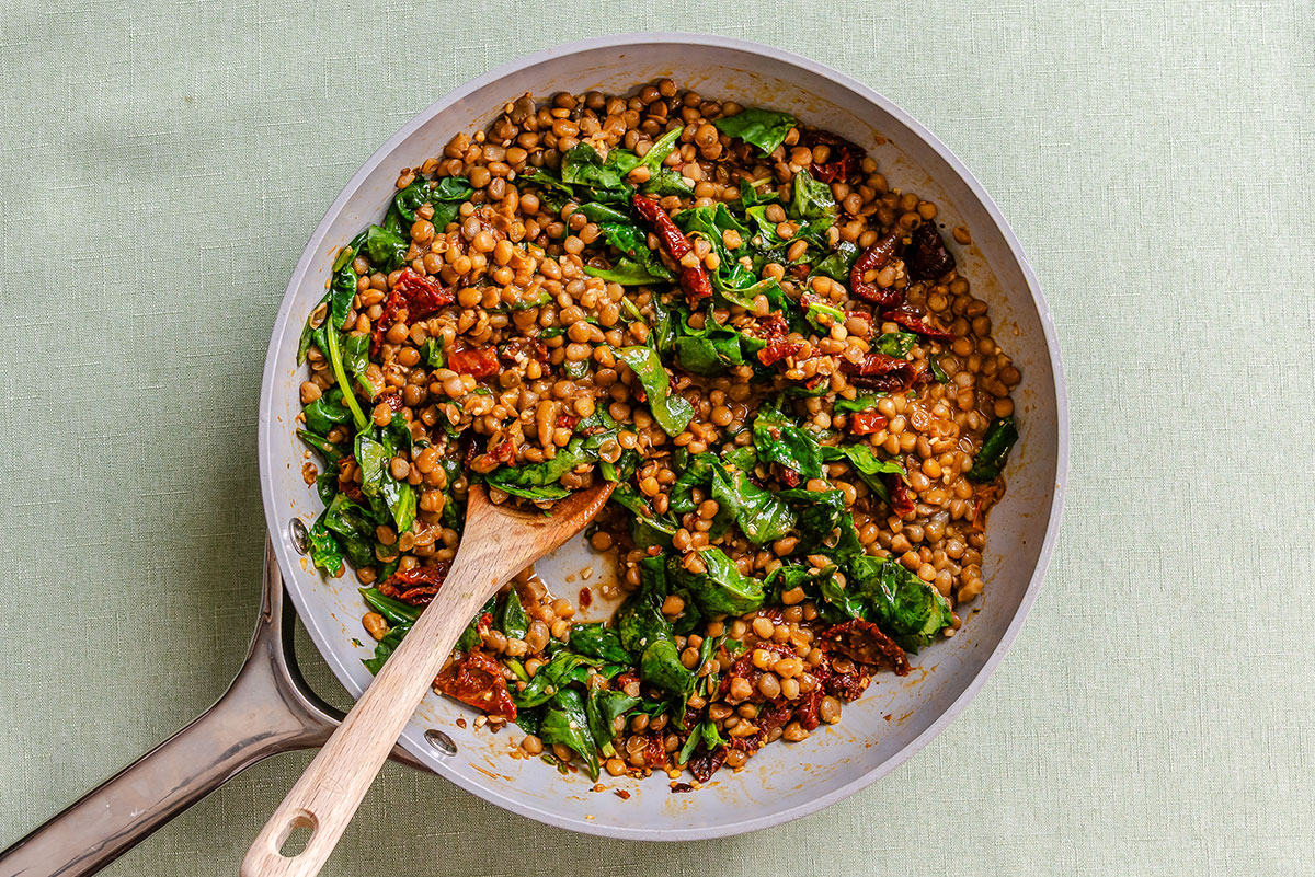 A skillet containing cooked lentils, spinach, and sun-dried tomatoes, with a wooden spoon resting in the pan.