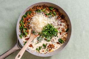A skillet containing lentils, spinach, and sun-dried tomatoes in a creamy sauce, topped with shredded cheese and fresh herbs, with a wooden spoon resting in the pan.